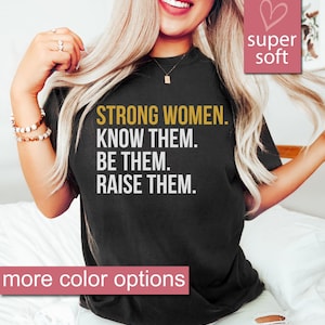 K&ouml;nnte beinhalten: Ein schwarzes T-Shirt mit dem Text "STRONG WOMEN. KNOW THEM. BE THEM. RAISE THEM." in goldener Schrift. Das Shirt wird von einer Person mit langen blonden Haaren modelliert. Der Text "super soft" und ein Herzsymbol befinden sich in der oberen rechten Ecke des Bildes. Der Text "more color options" befindet sich am unteren Rand des Bildes.
