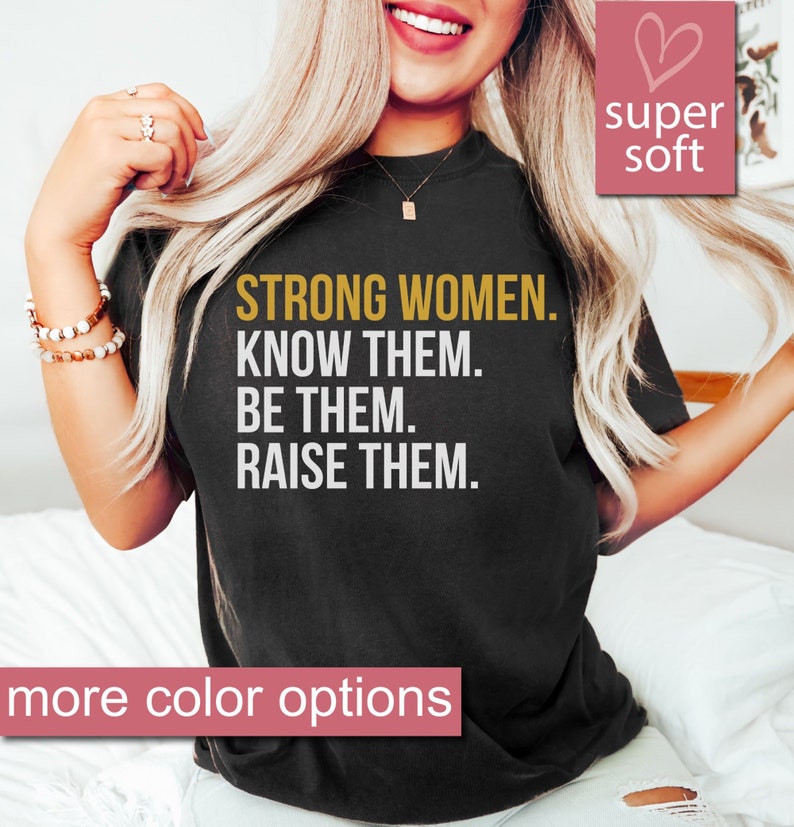 K&ouml;nnte beinhalten: Ein schwarzes T-Shirt mit dem Text "STRONG WOMEN. KNOW THEM. BE THEM. RAISE THEM." in goldener Schrift. Das Shirt wird von einer Person mit langen blonden Haaren modelliert. Der Text "super soft" und ein Herzsymbol befinden sich in der oberen rechten Ecke des Bildes. Der Text "more color options" befindet sich am unteren Rand des Bildes.