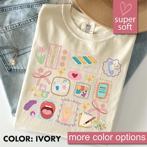 Könnte beinhalten: Elfenbeinfarbenes T-Shirt mit farbenfrohen grafischen Illustrationen. Das Design umfasst ein Xylophon, ein Telefon, ein Herz und das Wort "COMMUNICATION". Das Shirt ist mit "super soft" und "COMFORT COLORS" gekennzeichnet.