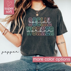 May include: A dark gray t-shirt with the text "SOCIAL WORKER" in rainbow colors. The text is outlined in black. The shirt is super soft and has a relaxed fit.