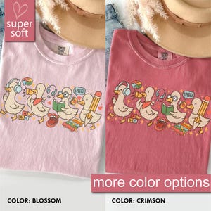 May include: Two t-shirts, one blossom pink and one crimson, featuring a cartoon duck design. The ducks wear headphones, read books, and hold pencils. The text "super soft" and "more color options" are also visible.