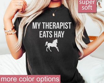 May include: A black t-shirt with the text "My Therapist Eats Hay" and a white silhouette of a horse. The shirt is super soft and has more color options.