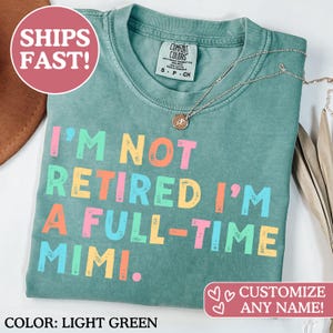 以下が含まれることがあります： 「I'M NOT RETIRED I'M A FULL-TIME MIMI」の文字がカラフルにプリントされた、ライトグリーンのTシャツ。シルバーのネックレスとペンダントがシャツの上に置かれています。「Comfort Colors」とラベルされ、サイズはSです。