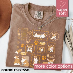 May include: Espresso-colored t-shirt featuring a repeating pattern of cartoon corgis in various poses, framed in gold. The shirt includes paw prints, dog bowls, and toy illustrations. The text "super soft" and "more color options" are also visible.