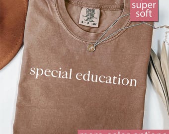 Puede incluir: Camiseta color espresso con las palabras "special education" en blanco. La camiseta tiene cuello redondo y una pequeña etiqueta en la parte superior de la espalda. Un collar plateado con un colgante está drapeado sobre la camiseta. Las palabras "super soft" están en un cuadro rosa.