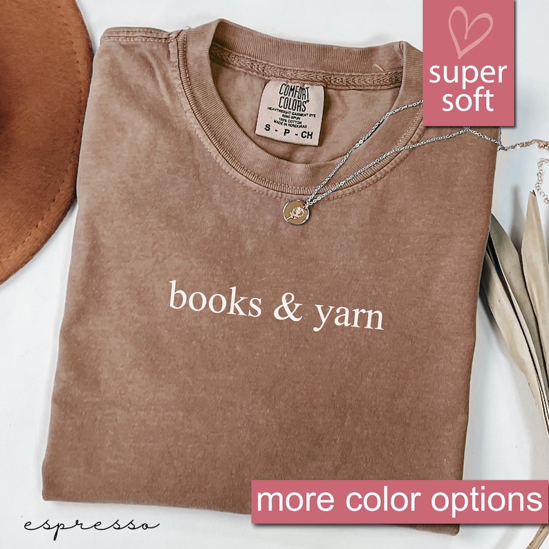 May include: Espresso-colored t-shirt with the words "books & yarn" in white. The shirt has a crew neck and a small tag. A silver necklace with a pendant rests on the shirt. The words "super soft" are in a pink box.