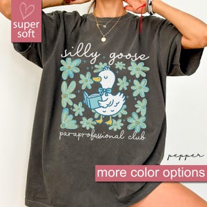 Puede incluir: Camiseta gris oscuro con un diseño caprichoso que presenta un ganso blanco leyendo un libro, rodeado de flores azules. El texto dice "silly goose + paraprofessional club". La camiseta también tiene las palabras "super soft" y "más opciones de color".