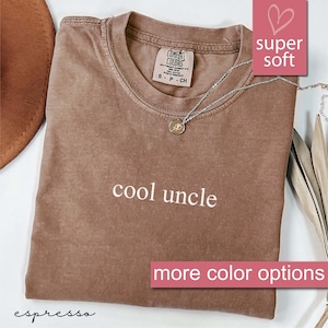 May include: A brown t-shirt with the text "cool uncle" printed on the front. The shirt is made of a soft, comfortable fabric and has a distressed look. The shirt is photographed on a white surface with a brown hat and a necklace.