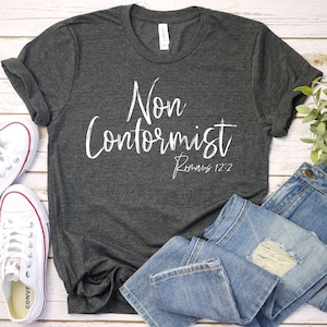 Puede incluir: Una camiseta gris oscuro con el texto "Non Conformist Romans 12:2" en letras blancas. La camiseta est&aacute; colocada sobre una superficie de madera con un par de zapatillas blancas y unos vaqueros azules.