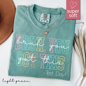 Può includere: T-shirt verde chiaro con la scritta "Bruh, You Got This - Test Day!" in lettere a blocchi colorate. La maglietta ha un girocollo e maniche corte. Una collana d'oro con un ciondolo è drappeggiata sulla maglietta.