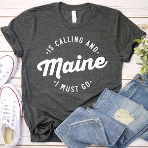 Może przedstawiać: Ciemnoszara koszulka z białym napisem "Is Calling and Maine I Must Go". Na zdjęciu niebieskie dżinsy i białe trampki.