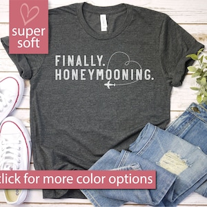 Puede incluir: Una camiseta gris oscuro con el texto "FINALLY. HONEYMOONING." en blanco. Un pequeño avión y un corazón también están impresos en la camiseta.