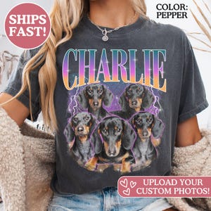 K&ouml;nnte beinhalten: Ein pfefferfarbenes T-Shirt mit einem Grafikdruck von f&uuml;nf schwarz-lohfarbenen Dackeln, die in einer Gruppe angeordnet sind. &Uuml;ber den Hunden ist der Name CHARLIE in einer gro&szlig;en Verlaufs-Schriftart gedruckt. Der Hintergrund enth&auml;lt violette Blitze.