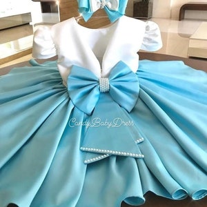 Op de afbeelding: Een lichtblauwe satijnen babyjurk met een wit lijfje en een grote blauwe strik met parel accenten. De jurk heeft een bolle rok en bijpassende polsbandjes met parels. De tekst "Candy Baby Dress" is zichtbaar op de rok.
