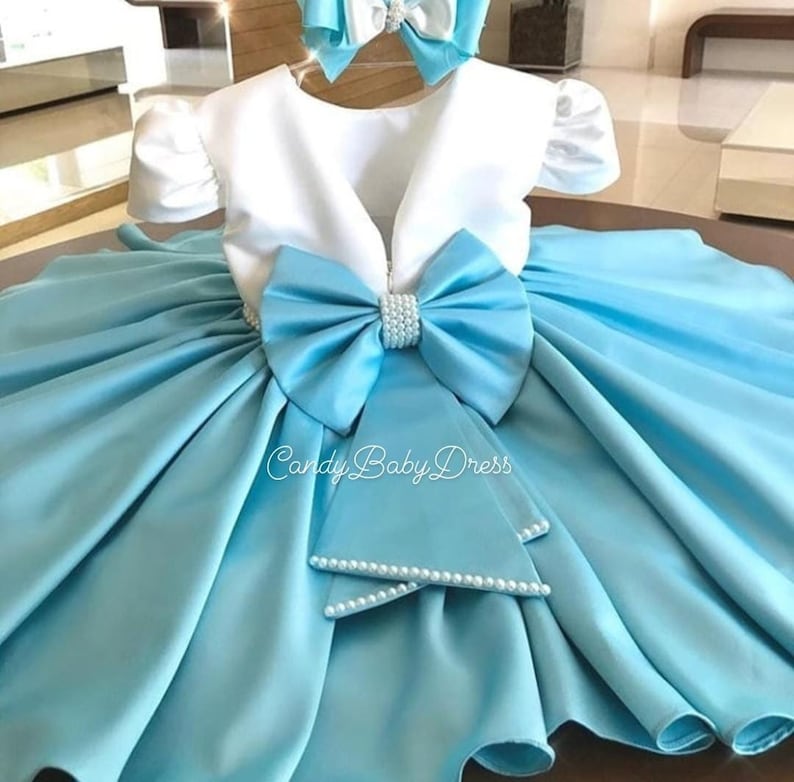 Op de afbeelding: Een lichtblauwe satijnen babyjurk met een wit lijfje en een grote blauwe strik met parel accenten. De jurk heeft een bolle rok en bijpassende polsbandjes met parels. De tekst "Candy Baby Dress" is zichtbaar op de rok.