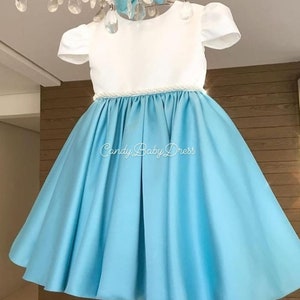 Op de afbeelding: Een lichtblauwe satijnen jurk met een wit bovenlijf en een parelrand rond de taille. De jurk heeft korte mouwen en een volle rok. De tekst "Candy Baby Dress" is op de rok gedrukt.