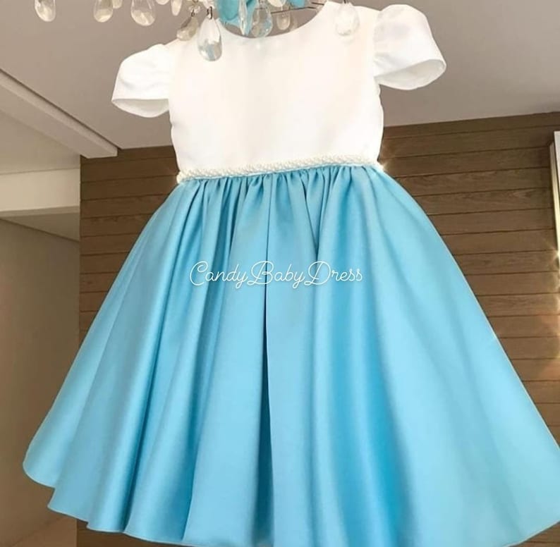 Op de afbeelding: Een lichtblauwe satijnen jurk met een wit bovenlijf en een parelrand rond de taille. De jurk heeft korte mouwen en een volle rok. De tekst "Candy Baby Dress" is op de rok gedrukt.