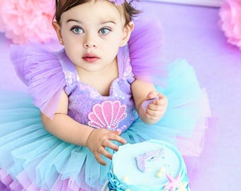 May include: A young child wearing a purple and blue tulle dress with a pink seashell design. The child is holding a blue and white frosted cake with a seashell design.