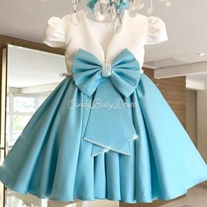 Op de afbeelding: Een lichtblauwe satijnen jurk met een wit bovenlijf en een grote blauwe strik met parel accenten. De jurk heeft een geplooide rok en een V-hals. De tekst "Candy Baby Dress" is zichtbaar op de rok.