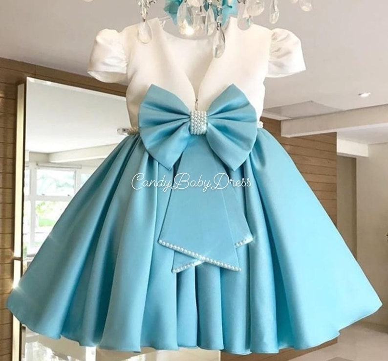 Op de afbeelding: Een lichtblauwe satijnen jurk met een wit bovenlijf en een grote blauwe strik met parel accenten. De jurk heeft een geplooide rok en een V-hals. De tekst "Candy Baby Dress" is zichtbaar op de rok.