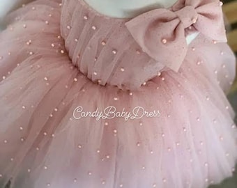 May include: A pink tulle dress with a large pink bow and pearl embellishments. The dress is on a white dress form. "Candy Baby Dress" is written on the skirt.