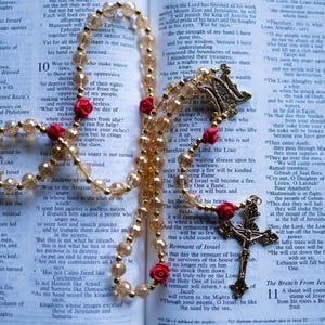 May include: A rosary featuring translucent gold-toned beads and small red rose-shaped accents, with a gold-toned metal crucifix and a central medal depicting a religious figure, resting on an open book with printed text in two columns.