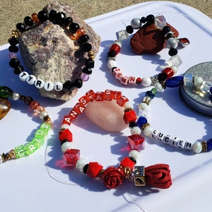 May include: Several beaded bracelets with names spelled out in letter beads. The bracelets feature a variety of beads in colors like black, red, white, and clear. Some bracelets have decorative elements like a red tassel and a rose.