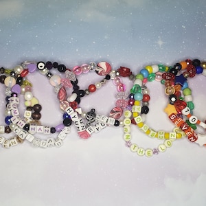 May include: A collection of beaded bracelets in various colors, featuring letter beads spelling out names and phrases. The bracelets include a mix of beads in white, brown, black, pink, and blue, with star and heart accents.