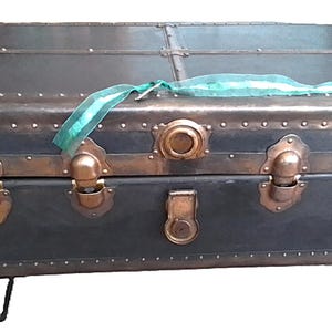 May include: A rectangular dark-colored trunk featuring metallic copper-toned corner reinforcements, trim, and latches. The exterior is accented with numerous small rivets along the edges. A translucent green ribbon rests across the top lid.
