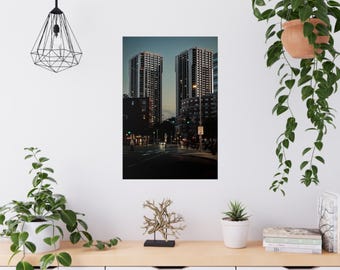 May include: A photograph of a city street at dusk, featuring two tall, modern buildings with illuminated windows. The street is lined with streetlights and traffic signals, with a few cars and pedestrians visible. The sky is a gradient of blue and gray.