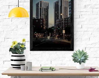 May include: A framed photograph of two tall buildings at dusk. The buildings are lit up with many windows. The photograph is in a black frame and is displayed on a wooden table. A yellow pendant lamp hangs above the table.