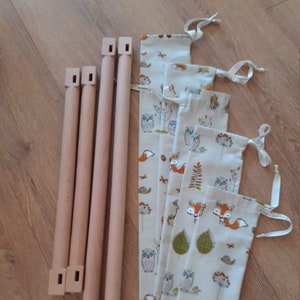 May include: Four wooden dowels with a square cut-out at each end. The dowels are arranged vertically and are next to five fabric pouches with a white background and a pattern of owls, foxes, and leaves in shades of brown, orange, and green.
