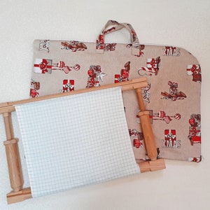May include: A wooden embroidery hoop with white fabric stretched across it. The hoop is on top of a beige fabric bag with a Christmas print featuring red and white gift boxes, ornaments, and the word "XMAS".