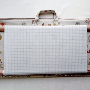 May include: A wooden embroidery hoop with a grid-patterned fabric stretched across it, set inside a patterned carrying case. The case features a repeating design of owls, foxes, and trees. The hoop has adjustable wooden screws and springs.