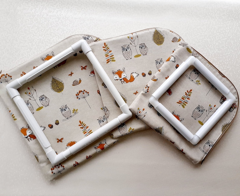 May include: Two white plastic embroidery hoops on a beige fabric with a woodland pattern of owls, foxes, and trees. The larger hoop is 10 inches in diameter and the smaller hoop is 6 inches in diameter.