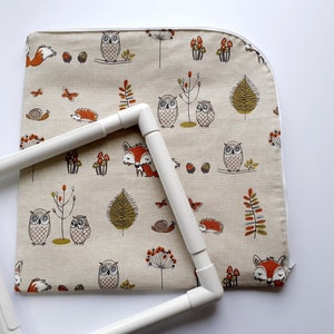 May include: A white embroidery hoop with a beige fabric inside. The fabric has a repeating pattern of owls, foxes, mushrooms, and other woodland creatures.