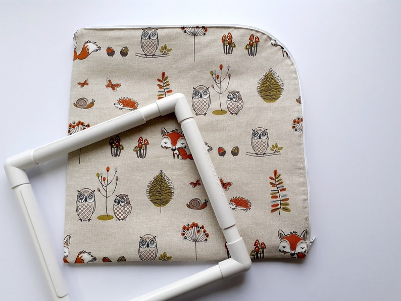 May include: A white embroidery hoop with a beige fabric inside. The fabric has a repeating pattern of owls, foxes, mushrooms, and other woodland creatures.