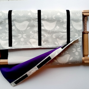 Peut inclure: Un cerceau à broder en bois avec un tissu gris à motif d'oiseau blanc et floral. Le tissu est fixé avec un ruban noir. Un tissu violet est partiellement enroulé autour du cerceau.