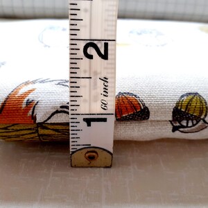 May include: A fabric with a whimsical fox and snail pattern in orange, yellow, and blue on a cream background. The fabric is approximately 1 inch thick, as measured by a 60-inch measuring tape.