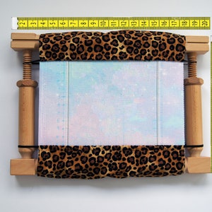 May include: A wooden embroidery hoop with a leopard print fabric cover. The fabric is stretched over a light blue and white patterned canvas. A yellow measuring tape is placed on top of the hoop, showing measurements in inches.