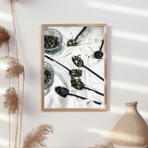 May include: A framed photograph of a white cloth with black spoons filled with dried herbs and flowers. The spoons are arranged in a fan shape. There is a small glass bowl filled with dried herbs and flowers in the upper left corner of the image.