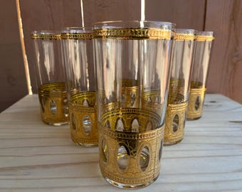 May include: Set of six vintage clear drinking glasses with gold-colored trim and decorative bands. The bands feature an arched design. The glasses are cylindrical and sit on a light-colored wooden surface.