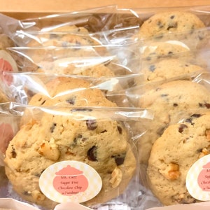 May include: A close-up of a variety of sugar-free chocolate chip cookies with walnuts, individually packaged in clear plastic bags. The cookies are labeled "Ms. Greez Sugar Free Chocolate Chip w/walnuts".