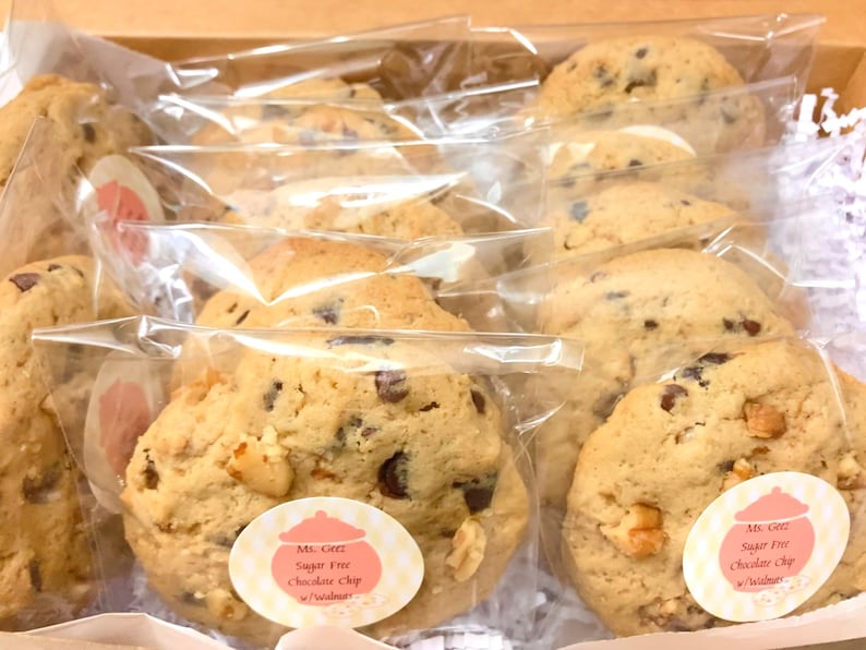 May include: A close-up of a variety of sugar-free chocolate chip cookies with walnuts, individually packaged in clear plastic bags. The cookies are labeled "Ms. Greez Sugar Free Chocolate Chip w/walnuts".