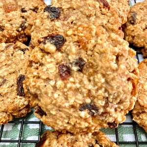 May include: A close-up of a stack of oatmeal raisin cookies on a wire rack. The cookies are golden brown and have a slightly crispy texture. They are studded with raisins and have a slightly sweet and nutty flavor.