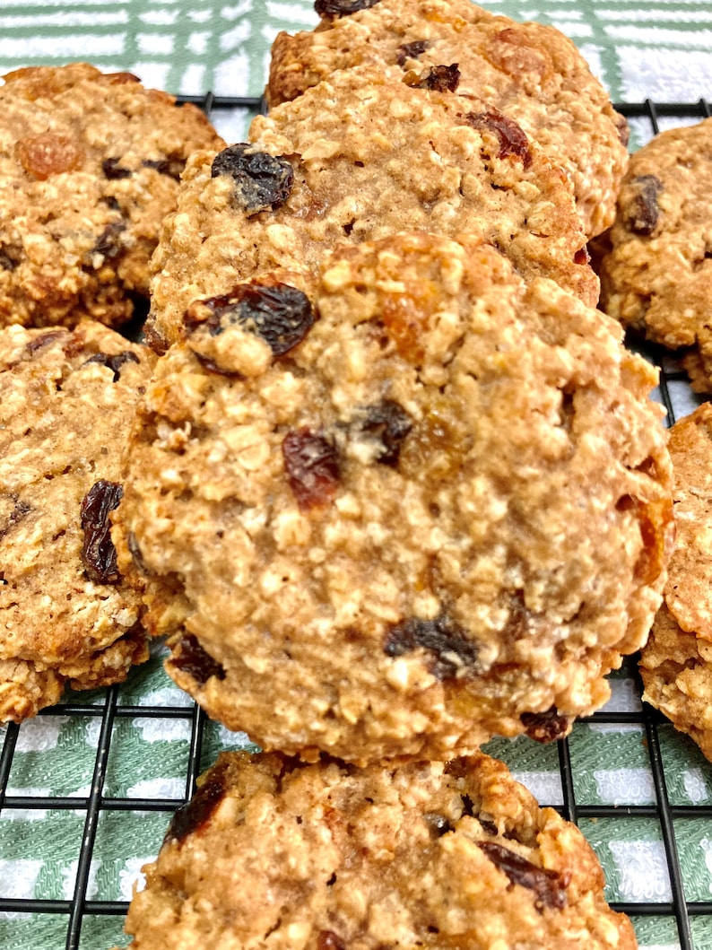 May include: A close-up of a stack of oatmeal raisin cookies on a wire rack. The cookies are golden brown and have a slightly crispy texture. They are studded with raisins and have a slightly sweet and nutty flavor.