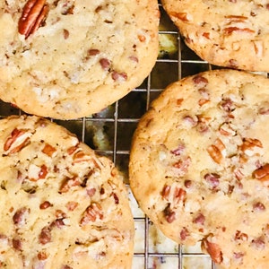 May include: Four golden brown chocolate chip cookies with pecans on a wire rack. The cookies are slightly domed and have a crispy texture.