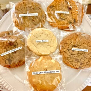 May include: Six different types of cookies in individual clear plastic bags on a white plate. The cookies are labeled: Oatmeal Raisin, Chocolate Chip Pecan, Dark Choc Chip Pecan, Sugar Cookie, White Choc Macadamia Nut, and Chocolate Chip.