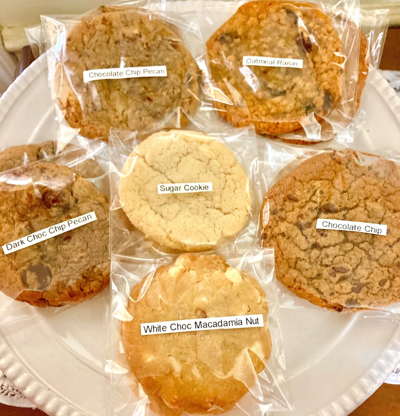May include: Six different types of cookies in individual clear plastic bags on a white plate. The cookies are labeled: Oatmeal Raisin, Chocolate Chip Pecan, Dark Choc Chip Pecan, Sugar Cookie, White Choc Macadamia Nut, and Chocolate Chip.