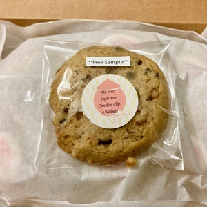 May include: A clear plastic bag containing a single sugar-free chocolate chip cookie with walnuts. The cookie has a pink and yellow label that reads "Ms. Geez Sugar Free Chocolate Chip w/Walnuts". The bag has a white label that reads "**Free Sample**".